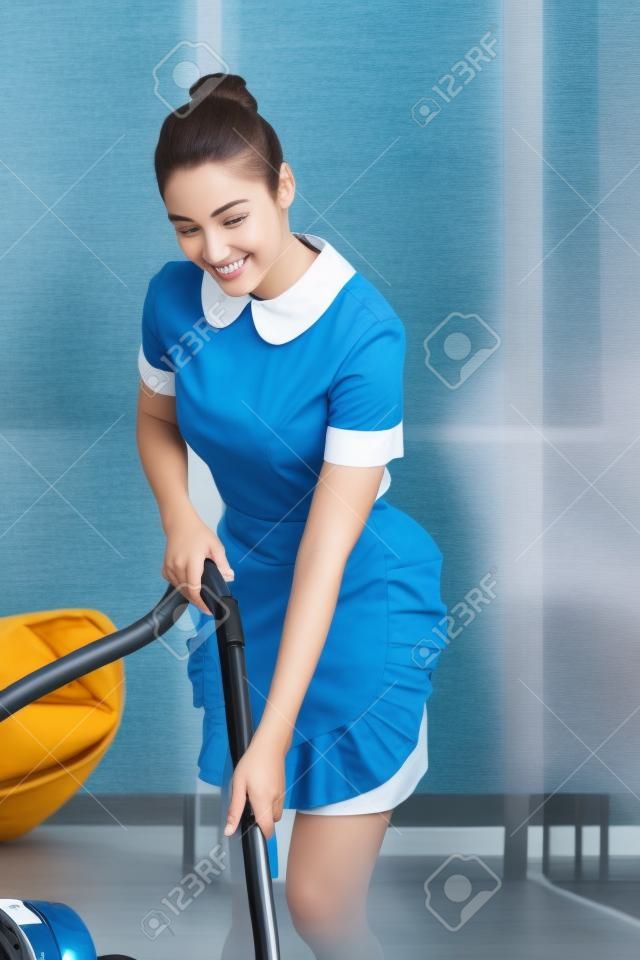 młoda uśmiechnięta pokojówka w mundurze za pomocą odkurzacza