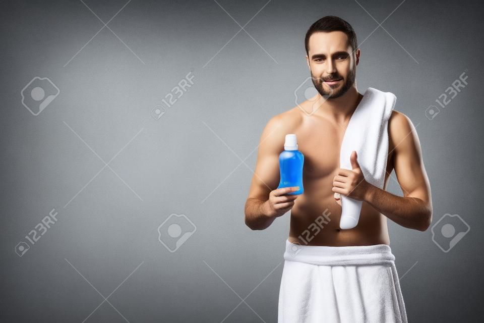 man with bath towel on shoulder holding bottle with tooth rinse in hand, isolated on gray