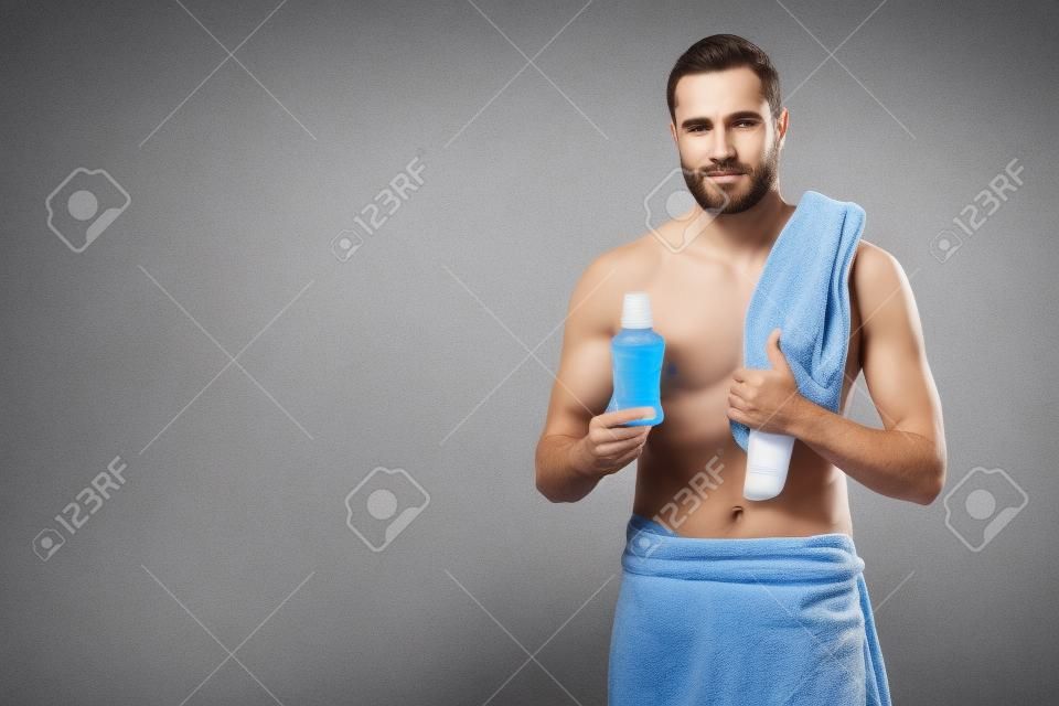 man with bath towel on shoulder holding bottle with tooth rinse in hand, isolated on gray