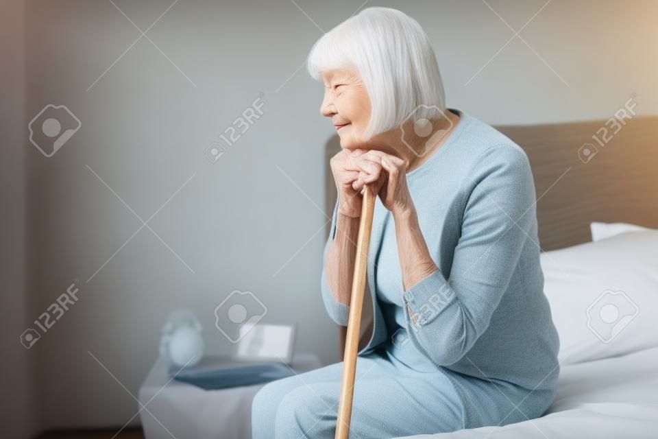 donna anziana sola con le mani sul bastone da passeggio seduta sul letto a casa