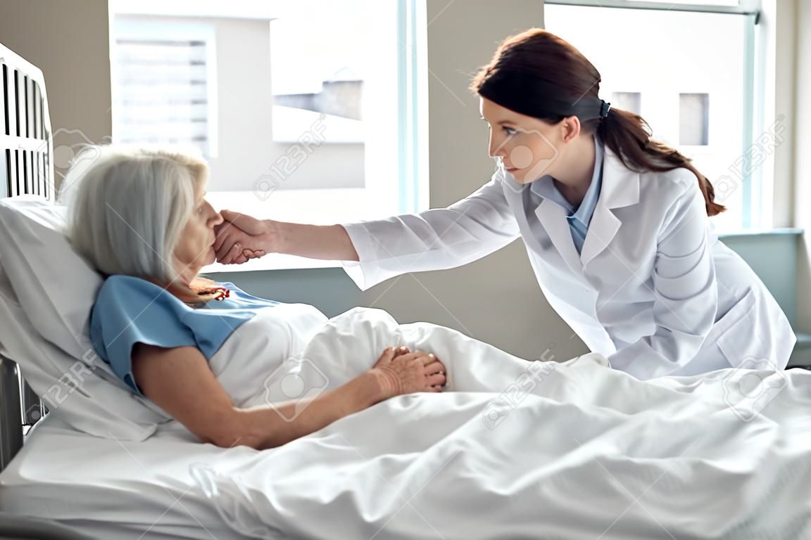 female doctor holding hands and supporting sad senior woman lying in hospital bed