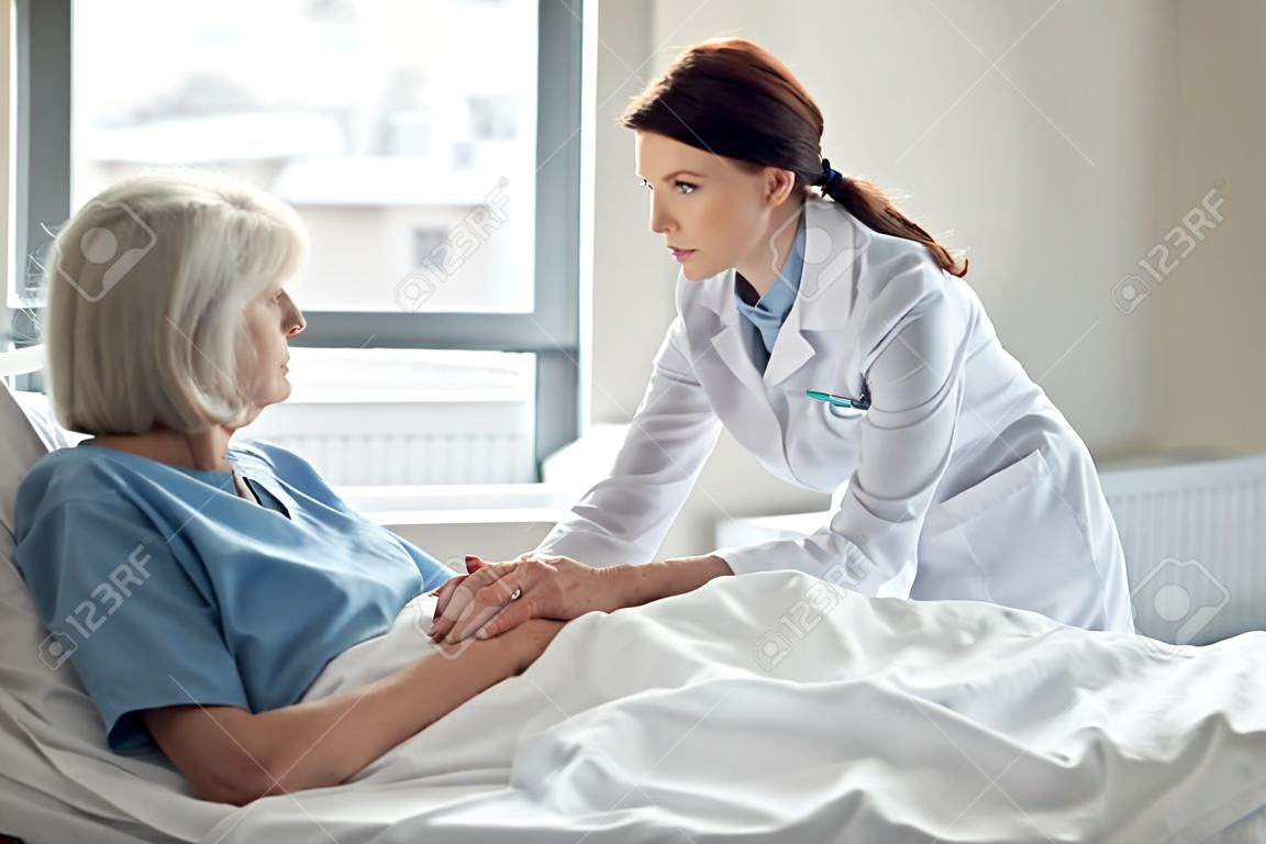 female doctor holding hands and supporting sad senior woman lying in hospital bed