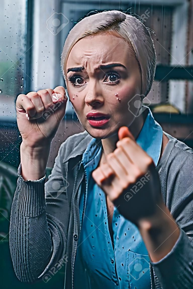 stressed adult woman with clenched fists looking at camera at home through window with raindrops