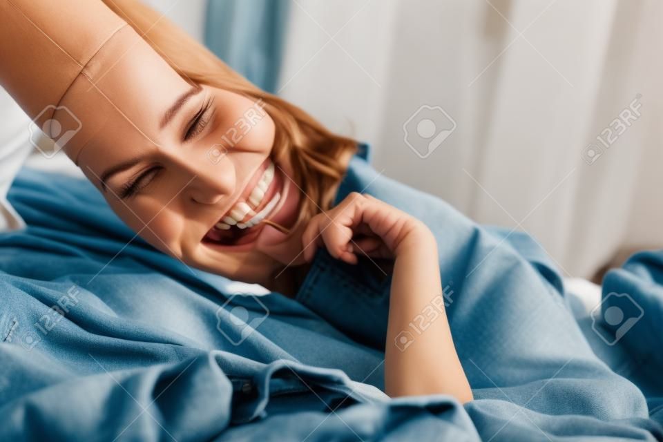 photo recadrée d'une jeune femme riant en mettant un pantalon en jean sur le lit
