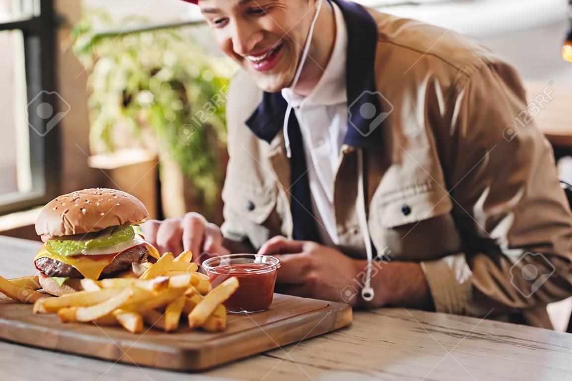 cropped view of cheerful man looking at tasty burger and french fries on cutting board in cafe