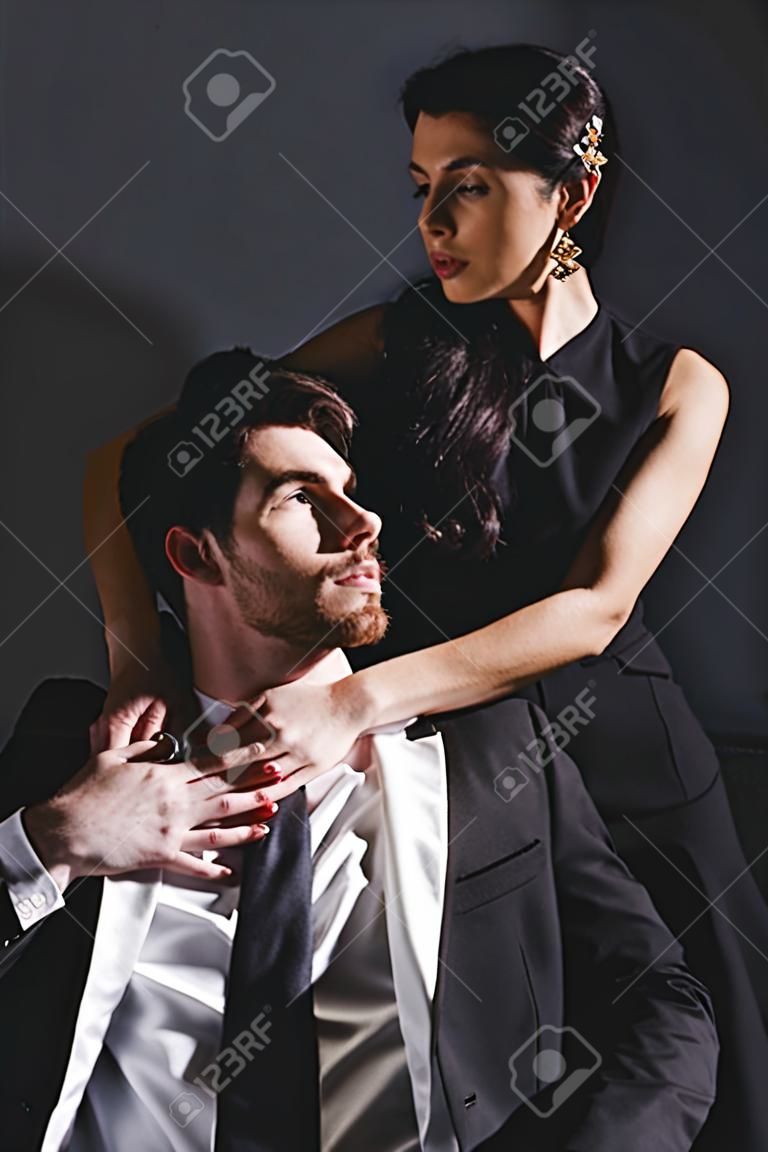 attractive woman in dress standing behind couch and hugging handsome man sitting on black background