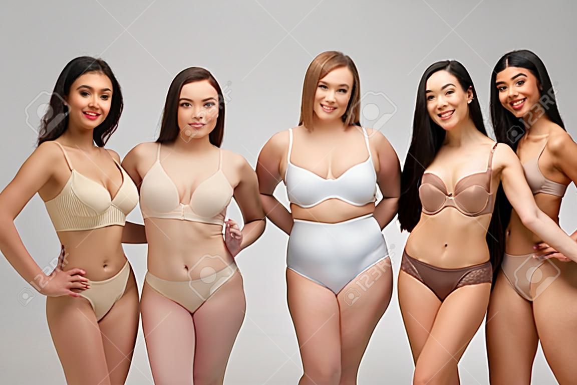 vijf mooie multiculturele meisjes in ondergoed kijken naar camera en glimlachen geïsoleerd op grijs, lichaam positiviteit concept