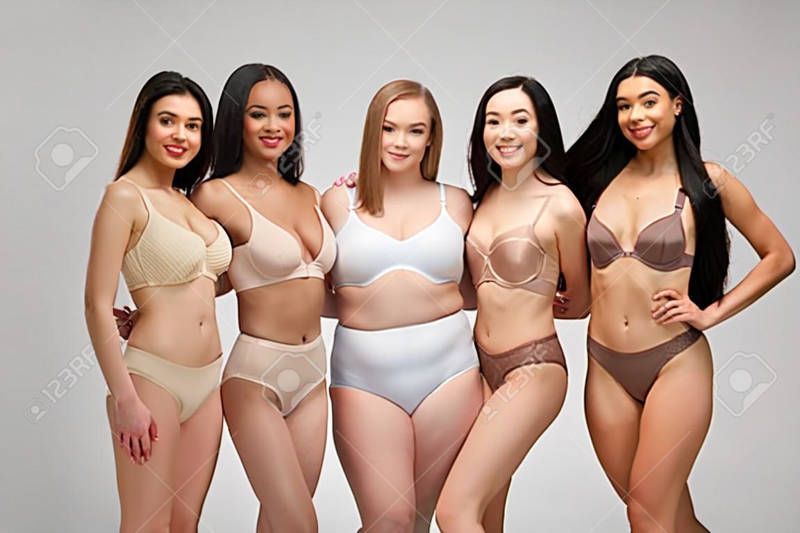 vijf mooie multiculturele meisjes in ondergoed kijken naar camera en glimlachen geïsoleerd op grijs, lichaam positiviteit concept