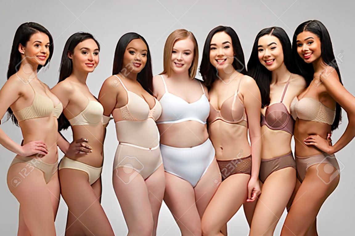 cinque belle ragazze multiculturali in biancheria intima che guardano la macchina fotografica e sorridono isolate su grigio, concetto di positività del corpo