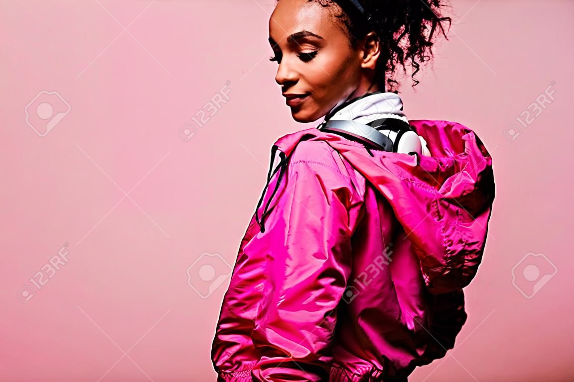 Hermosa deportista afroamericana en cazadora con auriculares aislado en rosa con espacio de copia