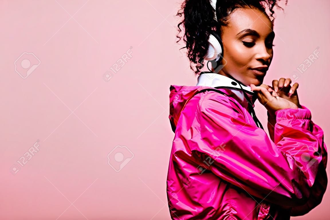 Piękna afroamerykańska sportsmenka w wiatrówce ze słuchawkami odizolowanymi na różowo z miejsca na kopię