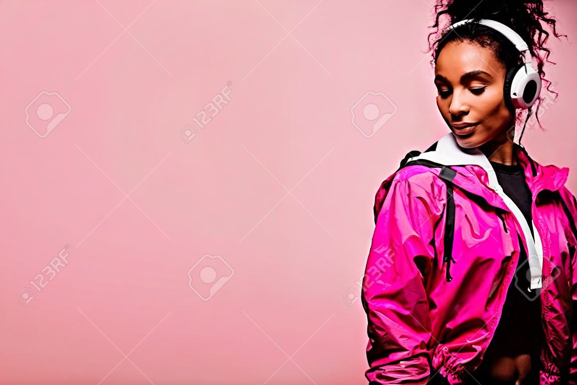 Piękna afroamerykańska sportsmenka w wiatrówce ze słuchawkami odizolowanymi na różowo z miejsca na kopię
