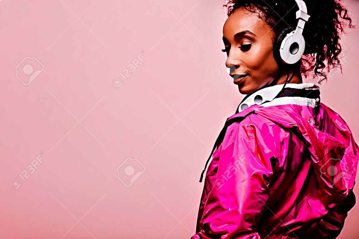 sportswoman americano africano bonito no windbreaker com fones de ouvido isolados no rosa com espaço da cópia