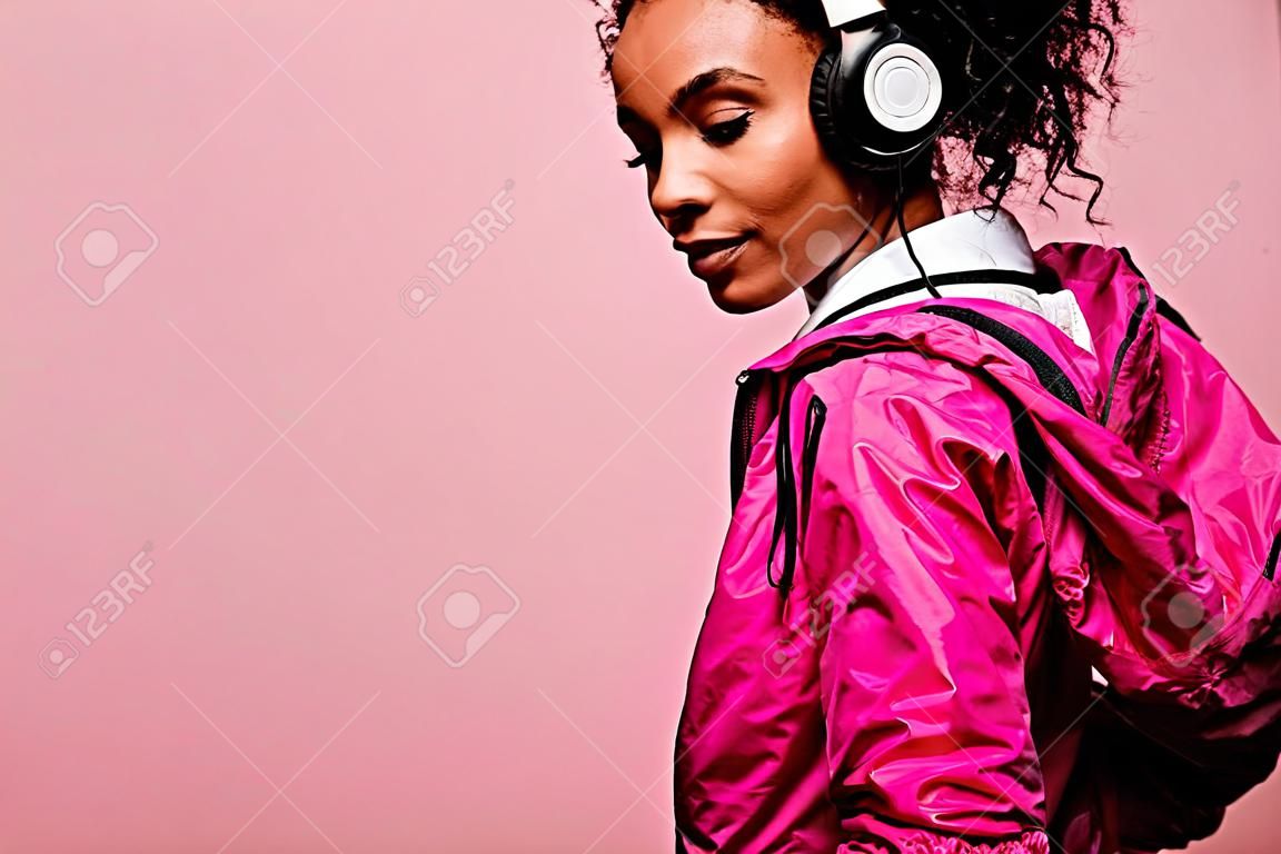 복사공간이 있는 분홍색으로 격리된 헤드폰을 끼고 윈드브레이커를 입은 아름다운 아프리카계 미국인 운동가