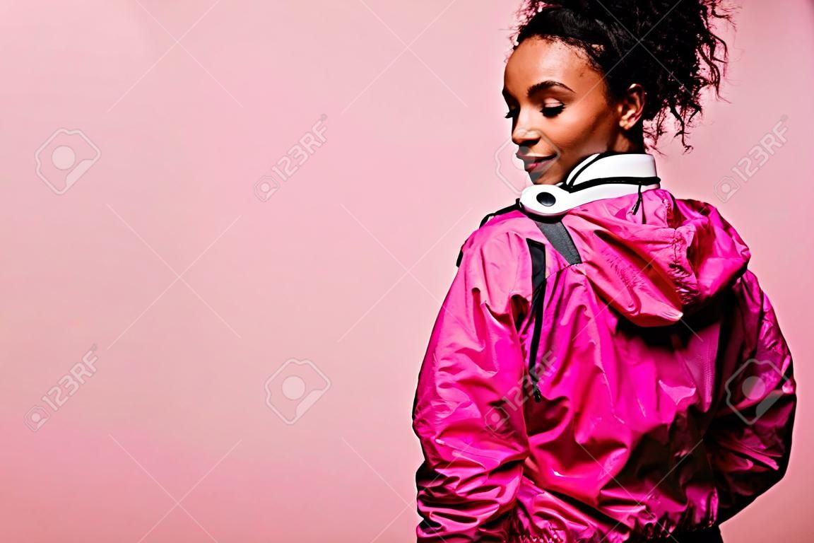 mooie Afrikaanse Amerikaanse sportvrouw in windbreaker met koptelefoon geïsoleerd op roze met kopieerruimte