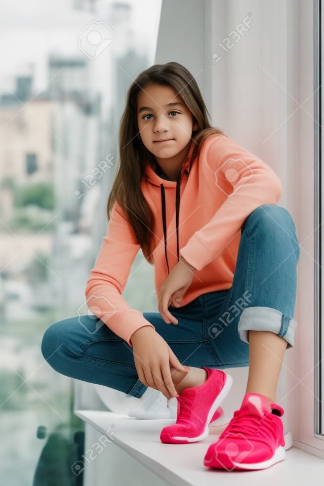 Curious teenager in sneakers sitting on window sill and looking at camera