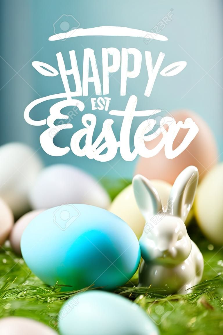 Oeuf peint en bleu près d'un lapin décoratif sur l'herbe verte avec des lettres de Joyeuses Pâques au-dessus