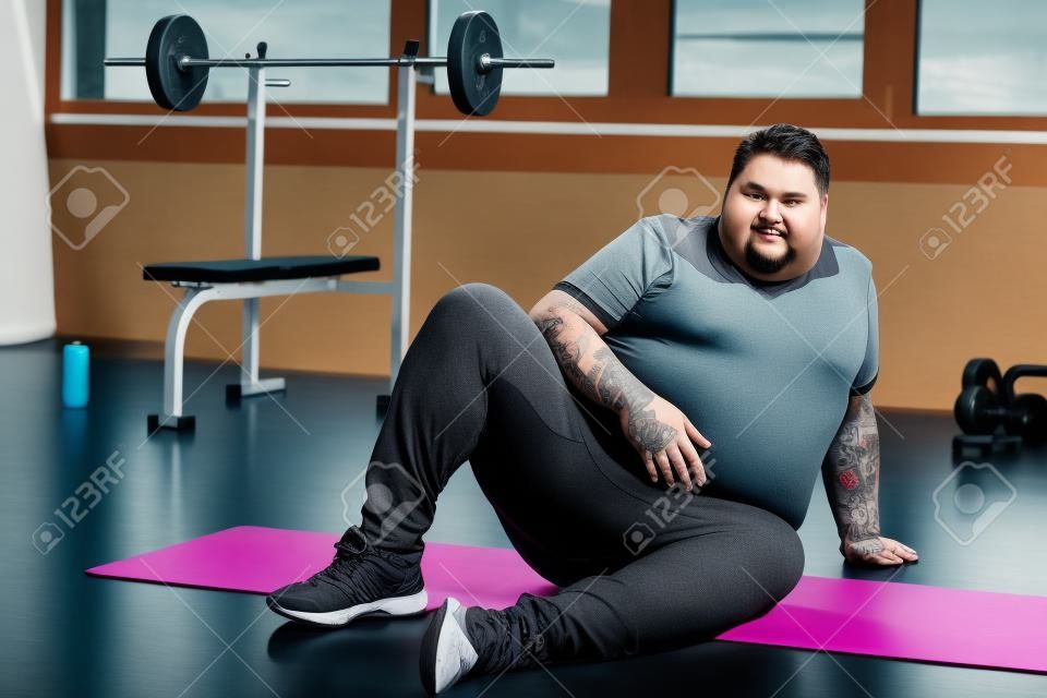 Hombre tatuado con sobrepeso mirando a la cámara y sentado en la colchoneta de fitness en el centro deportivo