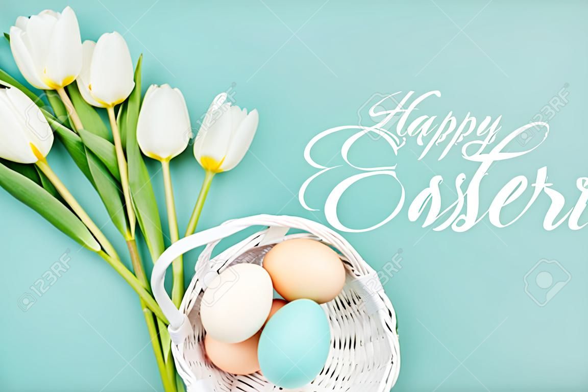 Vista dall'alto di uova di gallina dipinte in cesto di vimini e tulipani bianchi su sfondo blu con scritte bianche di buona Pasqua