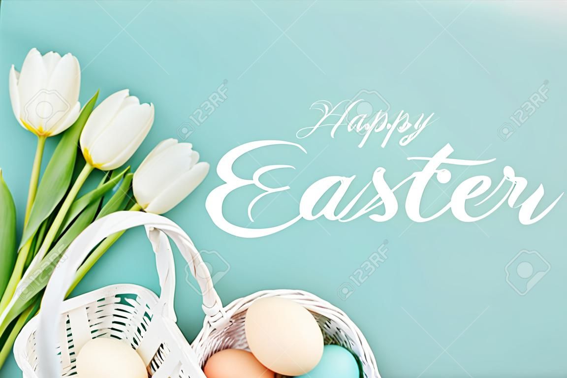 Vista dall'alto di uova di gallina dipinte in cesto di vimini e tulipani bianchi su sfondo blu con scritte bianche di buona Pasqua