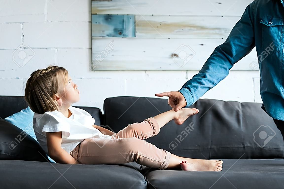 Cropped view of man pointing with finger at upset kid sitting on sofa at home