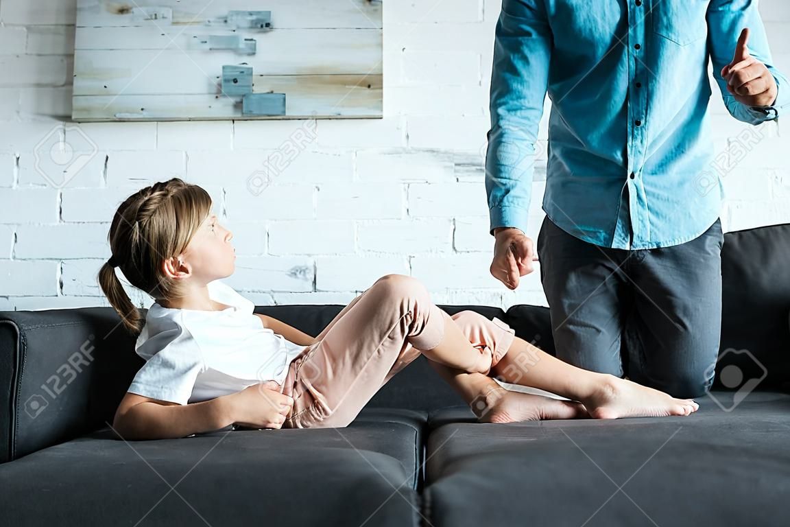 Vista recortada del hombre señalando con el dedo al niño molesto sentado en el sofá en casa