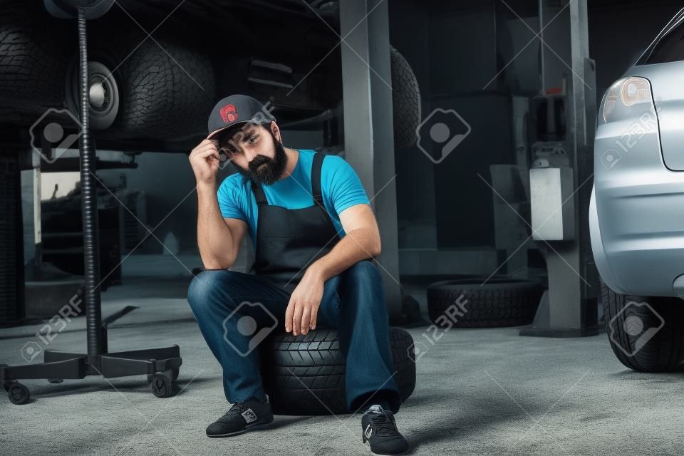 Upset bearded man touching cap and sitting on car tire