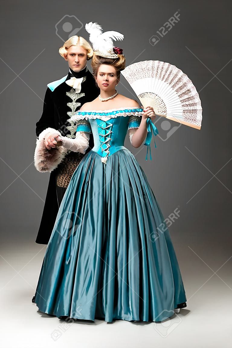jovem mulher vitoriana com ventilador de mãos dadas enquanto está de pé com o cavalheiro no cinza