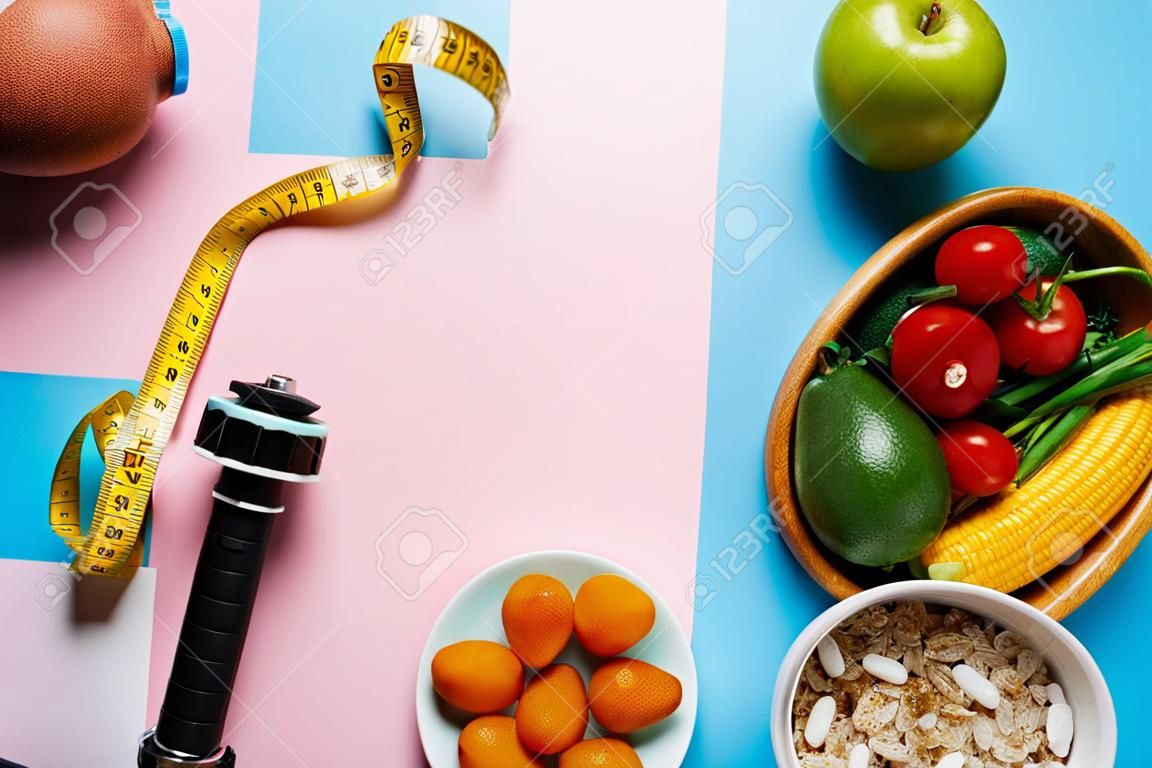 vista superior de deliciosa comida dietética y equipo deportivo con cinta métrica sobre fondo azul y rosa