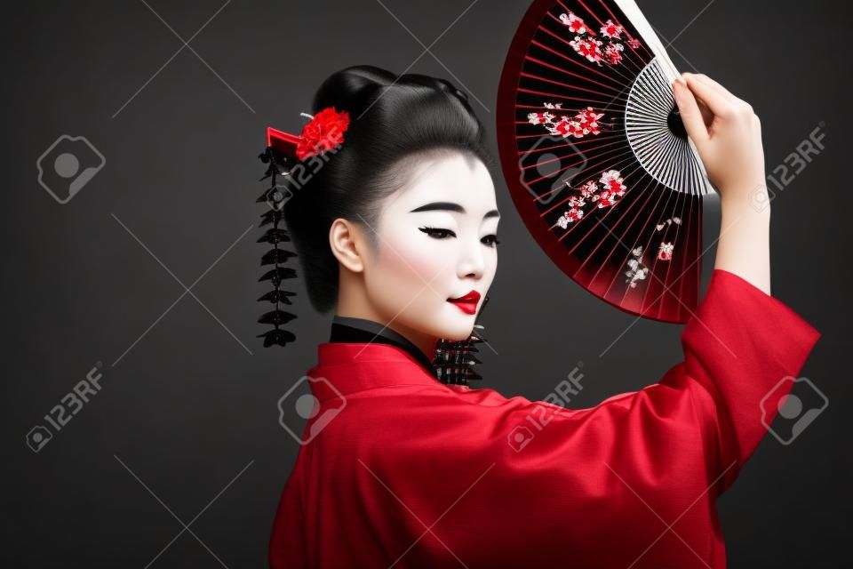 widok z boku gejszy w czarnym kimonie z czerwonymi kwiatami we włosach trzymających tradycyjny azjatycki wentylator ręczny odizolowany na czarno