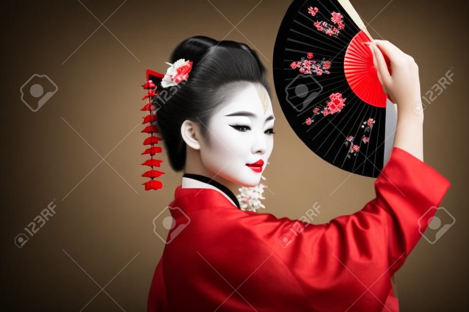 side view of geisha in black kimono with red flowers in hair holding traditional asian hand fan isolated on black