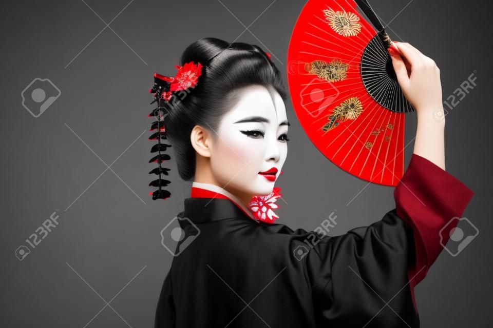 side view of geisha in black kimono with red flowers in hair holding traditional asian hand fan isolated on black