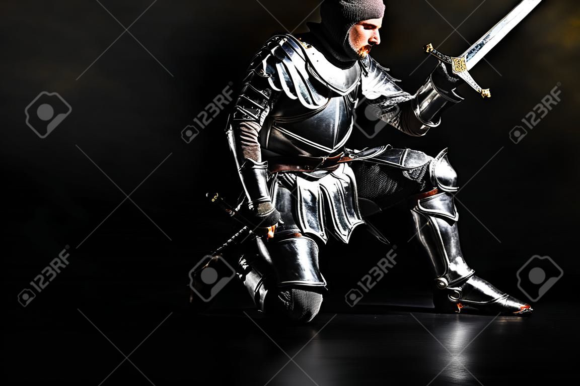 przystojny rycerz w zbroi trzymający miecz i zginający kolano na czarnym tle