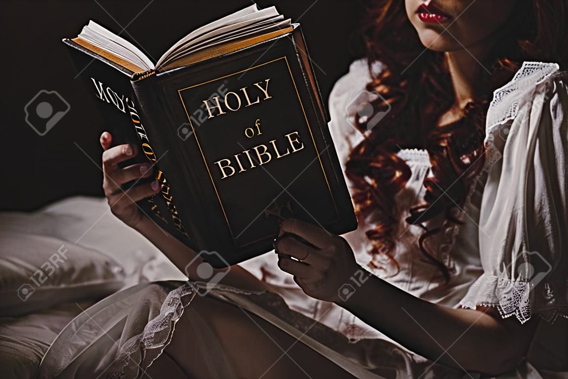 vista recortada del demonio femenino en camisón leyendo la sagrada biblia en la cama