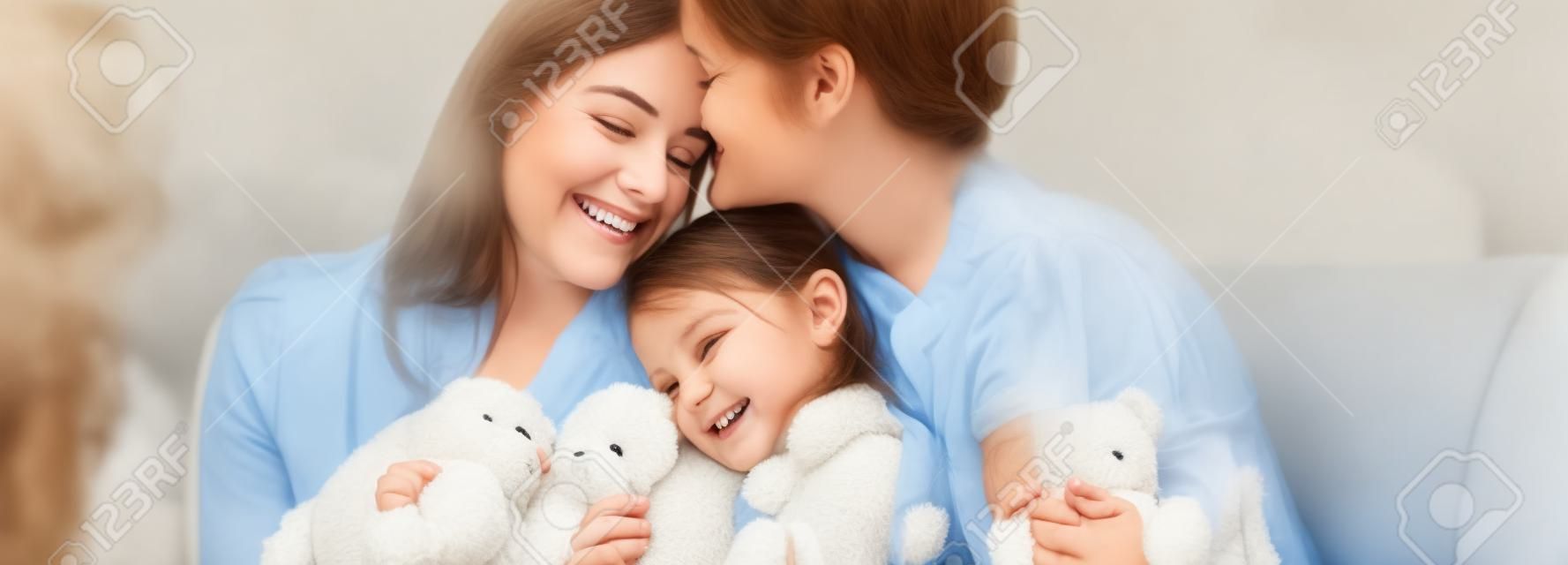 Madres felices abrazando a su hija sonriente con osito de peluche en el sofá, tiro panorámico