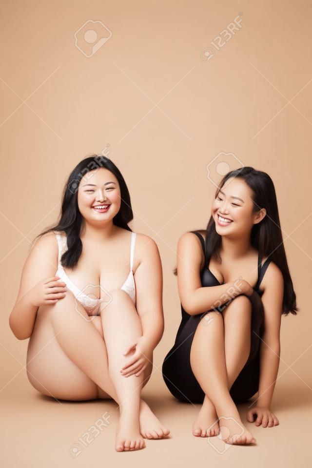 cheerful plus size girl and happy asian woman in lingerie sitting on beige, body positive concept
