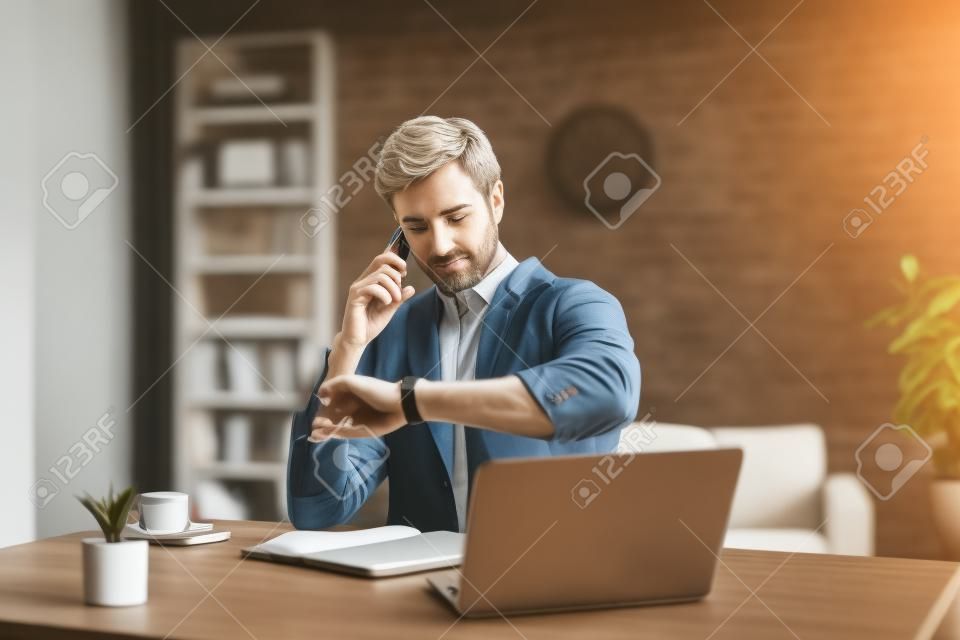 Selective focus of man checking time while talking on smartphone near laptop and notebook on table, concept of time management