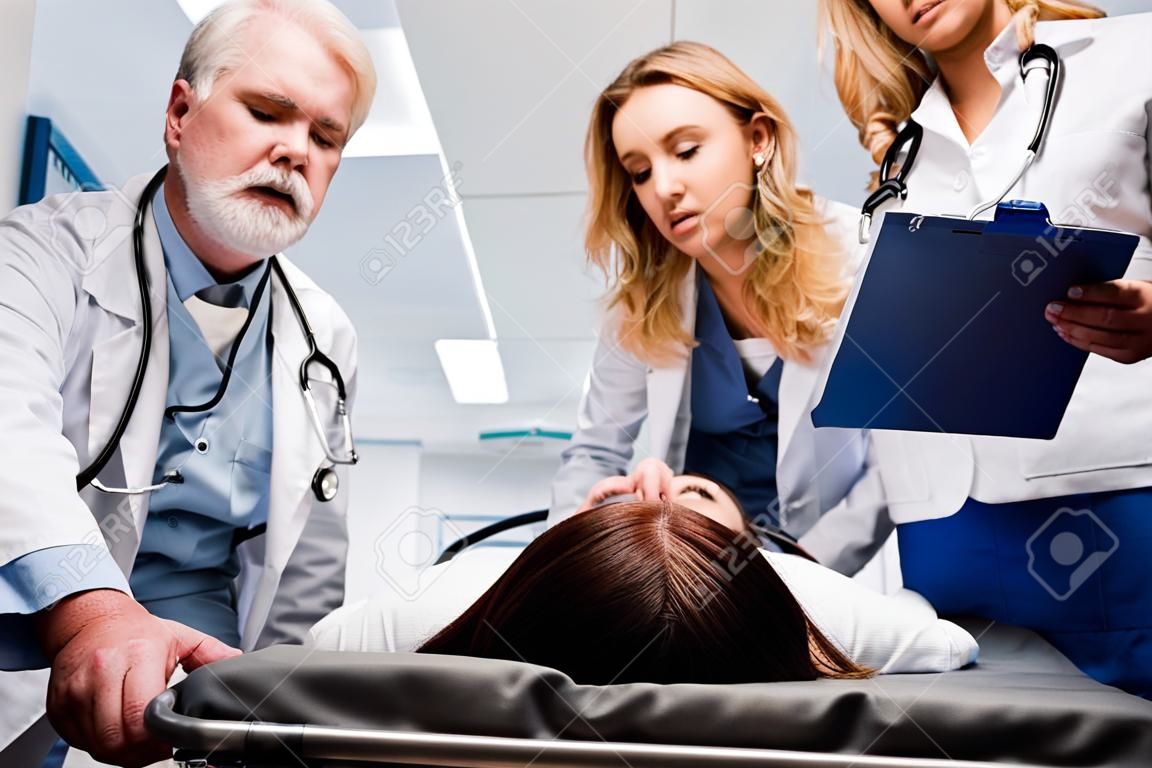 Enfoque selectivo del paciente acostado en camilla cerca de médicos con portapapeles en la clínica.
