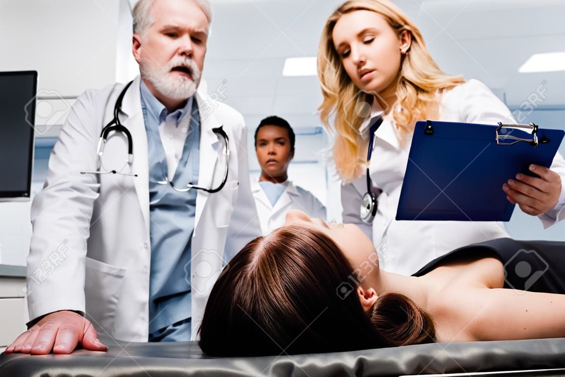 Enfoque selectivo del paciente acostado en camilla cerca de médicos con portapapeles en la clínica.