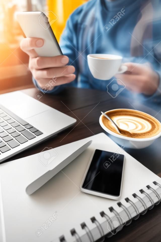 vista parziale del libero professionista che tiene tazza di caffè e smartphone vicino a laptop, notebook e carta di credito, sfondo sfocato