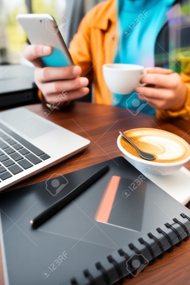 gedeeltelijk uitzicht van freelancer houden koffiebeker en smartphone in de buurt van laptop, notebook en creditcard, wazige achtergrond