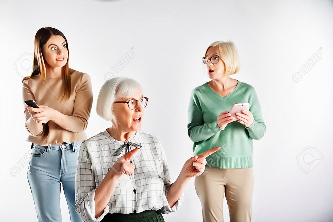 amazed senior woman in glasses holding smartphone and pointing with finger near daughter and granddaughter on blurred background