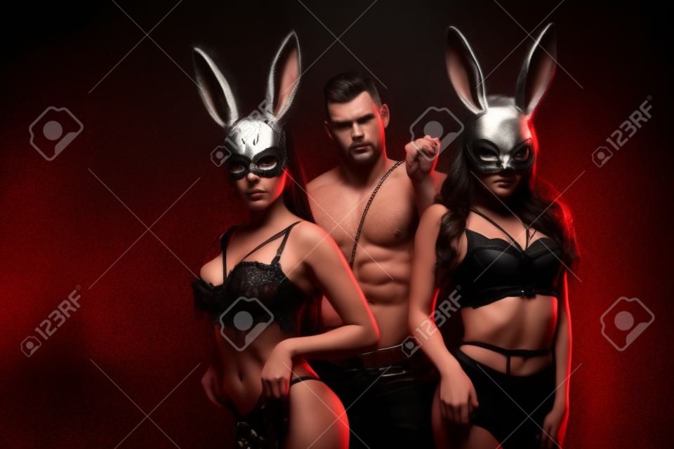 dominant man holding flogging whip near hot women in bunny masks on black background