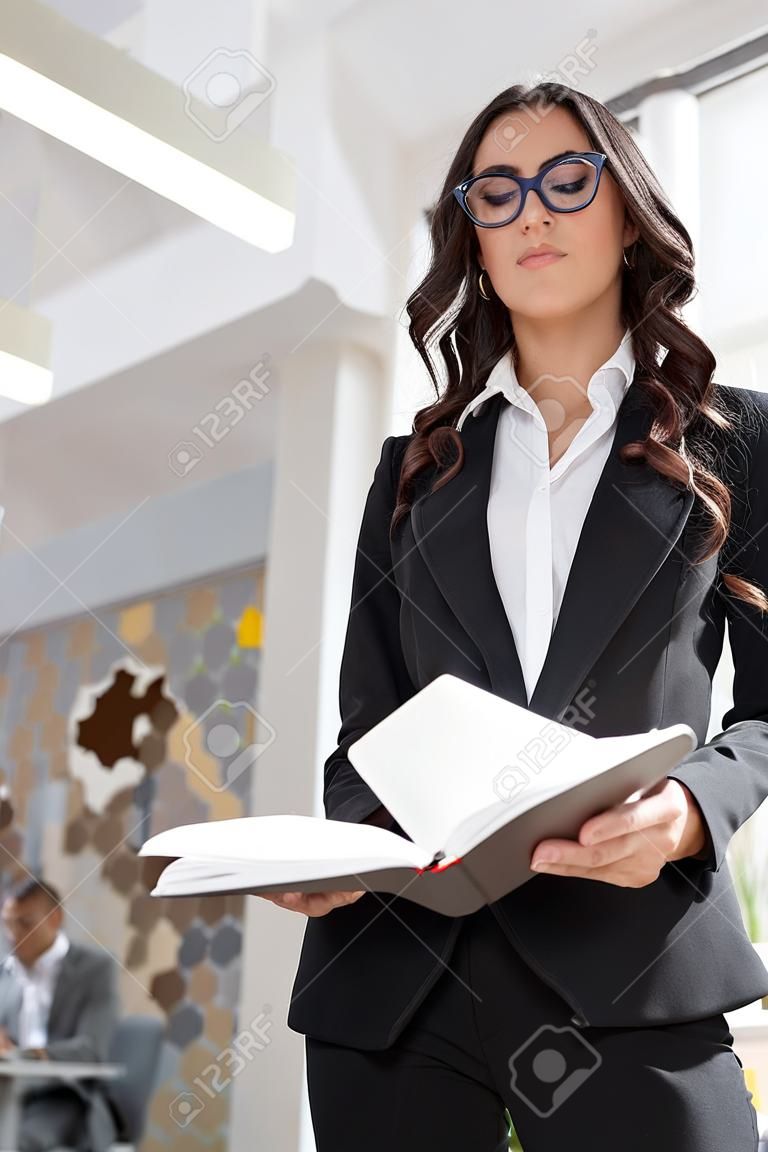 mooie brunette manager in bril en zwarte blazer houden pen en kijken in notebook in kantoor