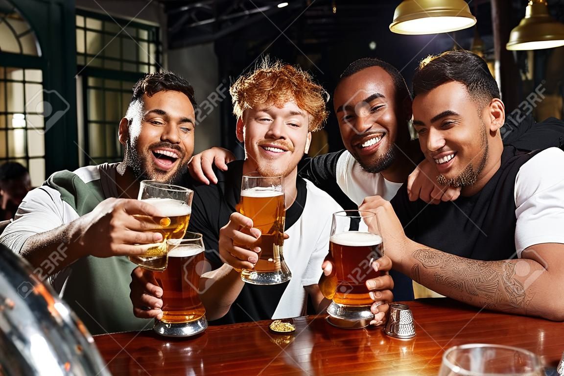 four happy multiethnic men holding glasses of beer during bachelor party, male friends in bar