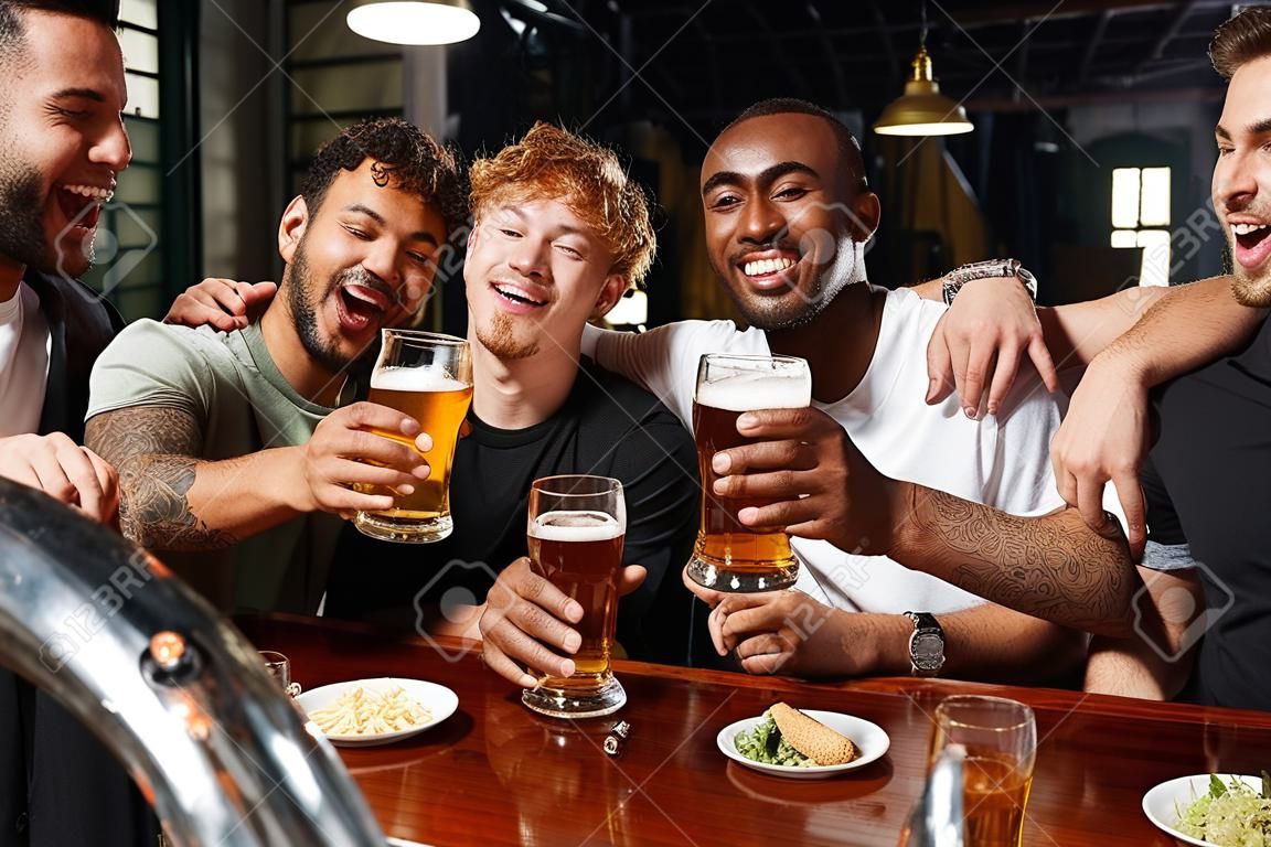 Cuatro hombres multiétnicos felices sosteniendo vasos de cerveza durante la despedida de soltero amigos varones en el bar