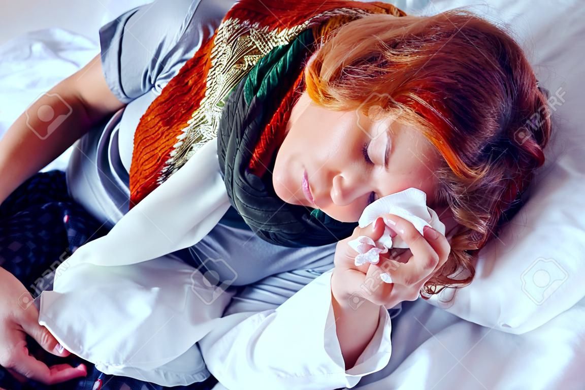 La donna esausta con una tazza di tè riposa a letto a causa della malattia. Una ragazza malsana con una sciarpa intorno al collo tiene un fazzoletto premuto sulla fronte.
