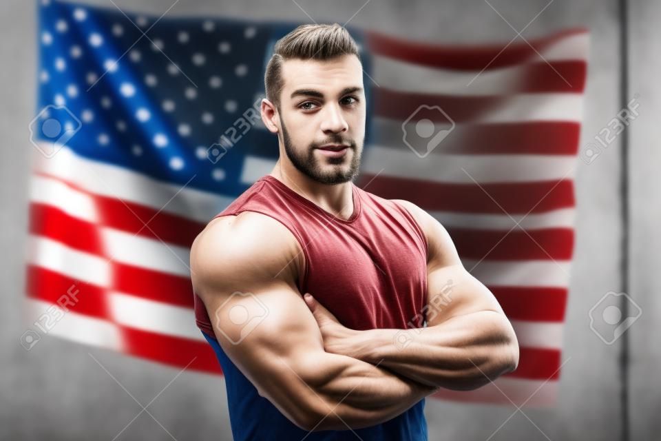 Patriotic American With Tattoo On His Hand Standing On The American Flag Background. The Successful Attractive Man Sported Physique In A White T-Shirt Looking Confident