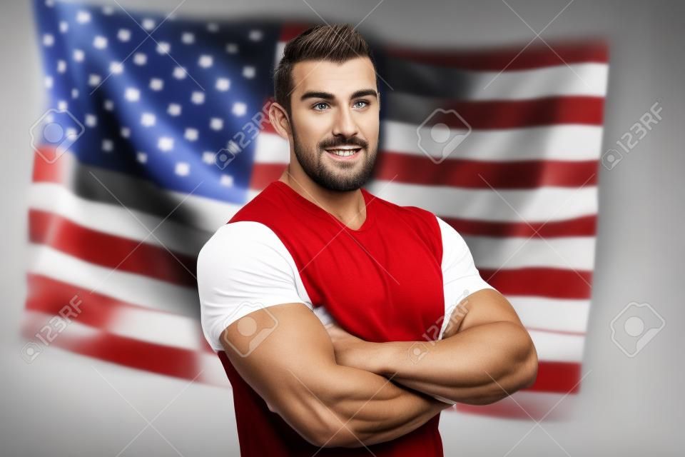 Patriotic American With Tattoo On His Hand Standing On The American Flag Background. The Successful Attractive Man Sported Physique In A White T-Shirt Looking Confident
