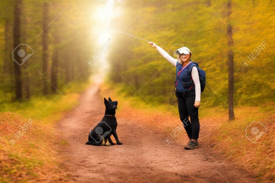 A Woman In Casual Training While Hiking On A Forest Road On A Warm Autumn Day. Black Purebred Shepherd Dog Sitting Fulfilling The Command Of The Hostess